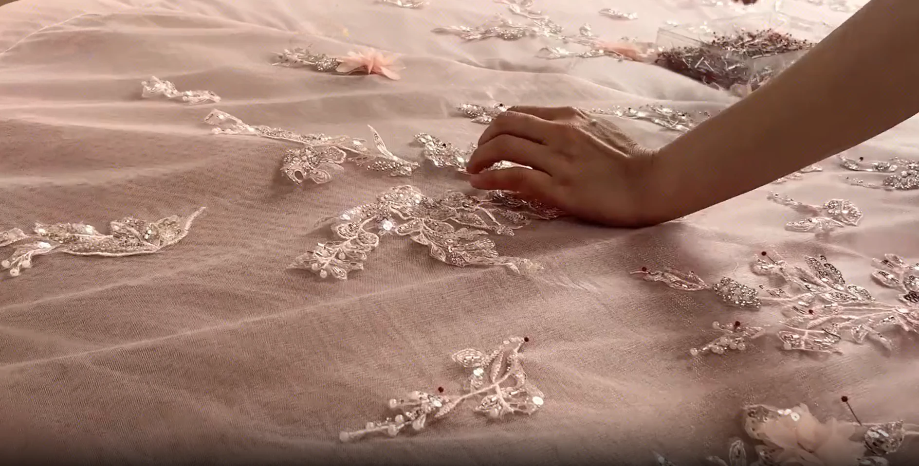 Hands positioning floral lace motifs across sheer pink tulle for a couture gown