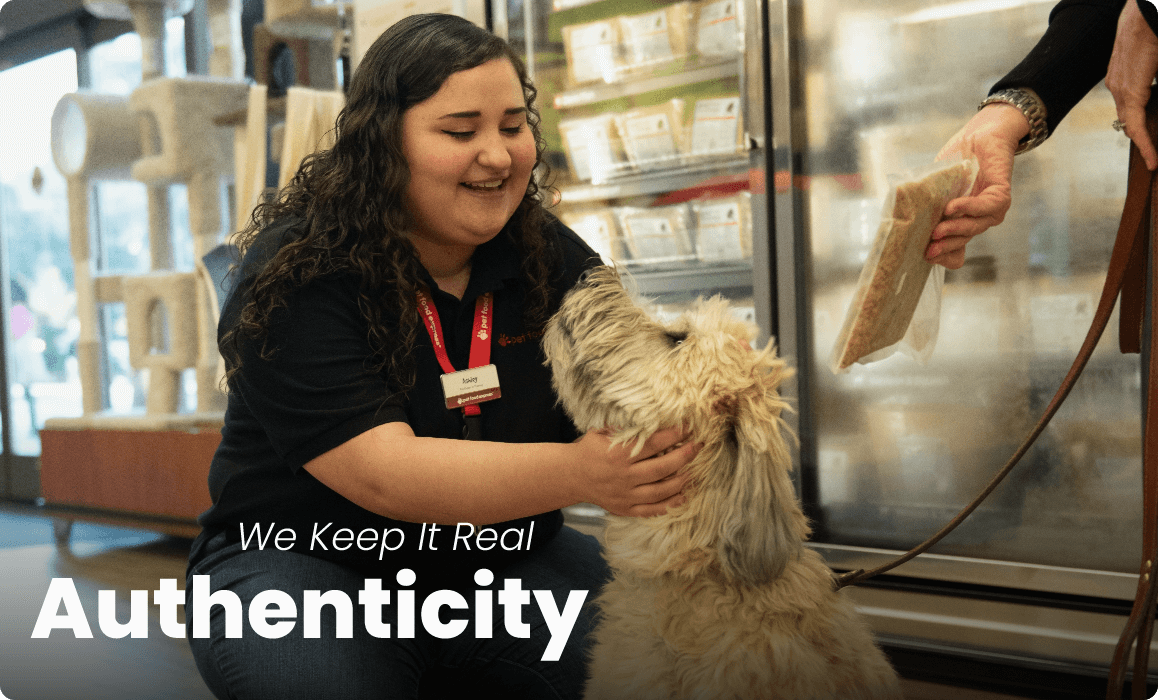 A Pet Food Express employee crouching to pet a dog. Text reads: We Kee It Real - Authenticity