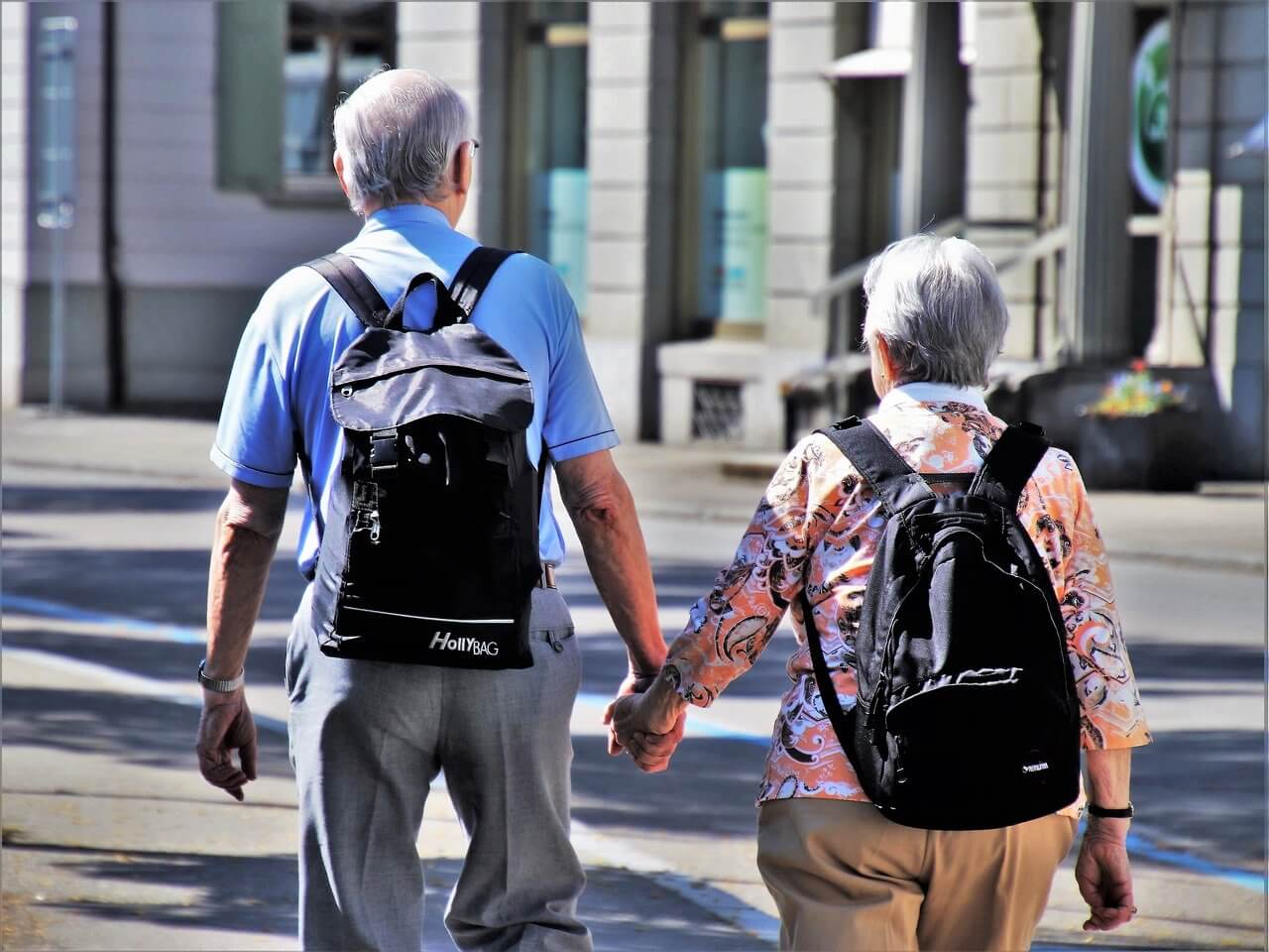 An elderly couple with backpacks holding hands and walking along an urban street in the daytime