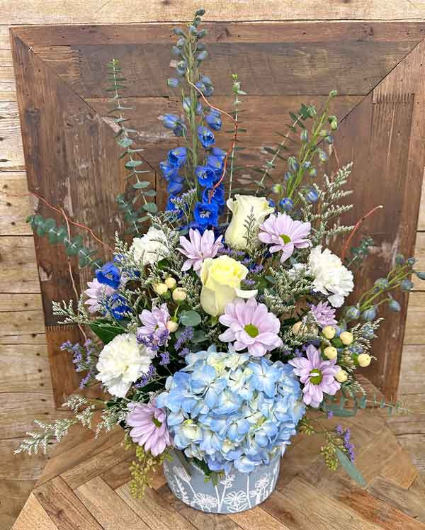 Blue and white flower arrangement with hydrangea roses delphinium and lavender daisies in decorative container