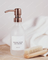 Flacon distributeur en verre givré à pompe cuivrée, étiquette Savon pour les mains, posé sur du bois près d'une brosse à ongles et d'une serviette