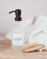 Flacon distributeur en verre givré à pompe noire, étiquette Savon pour les mains, posé sur du bois près d'une brosse à ongles et d'une serviette