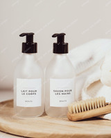 Deux flacons en verre givré à pompes noires, étiquettes Lait pour le corps et Savon pour les mains, sur un plateau rond en bois avec une brosse