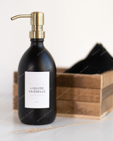 Flacon distributeur en verre noir mat à pompe laiton doré, étiquette Liquide vaisselle, devant une cagette en bois et un linge noir
