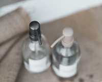 Vue plongeante et floue de deux distributeurs de savon en verre transparent pour la cuisine, pompes gris anthracite et beige, sur une toile de jute