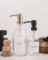 Deux flacons distributeurs en verre transparent, pompes laiton et noire, étiquettes Liquide vaisselle et Savon pour les mains, brosse en bois