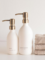 Deux flacons distributeurs blancs à pompes laiton, étiquettes Savon et liquide vaisselle mention kitchen's basics, devant des serviettes taupe