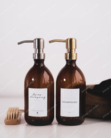 Deux distributeurs de savon en verre ambré côte à côte, l'un à pompe inox, l'autre à pompe dorée, étiquettes Après-shampoing et Shampoing