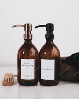Deux distributeurs de savon en verre ambré, l'un à pompe cuivrée, l'autre à pompe noire, étiquettes Après-shampoing et Shampoing, brosse en bois