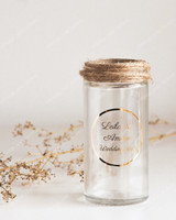 Flacon en verre transparent au col entouré de ficelle, sticker rond doré Leila & Amine Wedding day, près de fleurs séchées