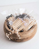 Sachets d'allumettes à têtes blanches, lilas et noires, empilés dans une coupelle en bois clair avec une branche de gypsophile séchée