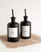 Deux bouteilles noires à bec verseur noir, étiquettes Huile blanche et Vinaigre balsamique, posées sur une toile de jute
