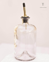 Bouteille en verre transparent équipée d'un bec verseur doré à embout noir, sur un linge blanc et fond beige