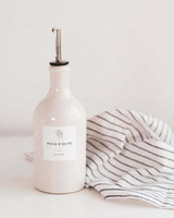 Bouteille huilier en grès beige équipée d'un bec verseur argenté, étiquette Huile d'olive, à côté d'un torchon rayé blanc et noir