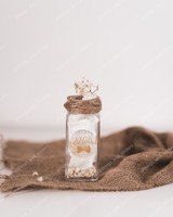 Sticker de naissance rond doré pailleté Nahil sur un petit flacon en verre garni de papier de soie, ficelle et gypsophile, sur une toile de jute