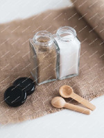 Deux pots à épices carrés ouverts sur une toile de jute, avec deux couvercles noirs et deux petites cuillères en bois posés à côté