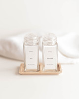 Deux pots à épices carrés à couvercles saupoudreurs transparents, étiquettes Sel et Poivre, dans un support en bambou devant un linge blanc