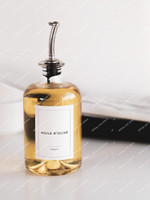 Huilier en verre transparent rempli d'huile, bec verseur argenté, étiquette Huile d'olive Basics, devant un linge blanc et un plateau noir