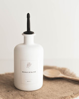 Bouteille ronde en verre blanc à bec verseur entièrement noir, étiquette Huile d'olive Basics, posée sur un set en jute