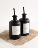 Deux huiliers noirs à bec verseur noir, étiquettes Huile blanche et Vinaigre balsamique, vus de trois quarts sur une toile de jute