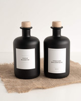 Deux huiliers ronds en verre noir à bouchons en bois, étiquettes Huile blanche et Vinaigre balsamique, posés sur de la jute