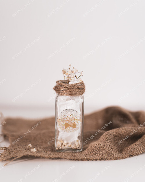 Sticker de naissance rond doré pailleté Nahil sur un petit flacon en verre garni de papier de soie, ficelle et gypsophile, sur une toile de jute