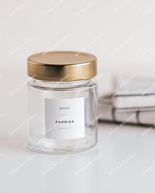 Pot à épices en verre à couvercle métal doré, étiquette blanche Epices Paprika, devant une pile de torchons rayés
