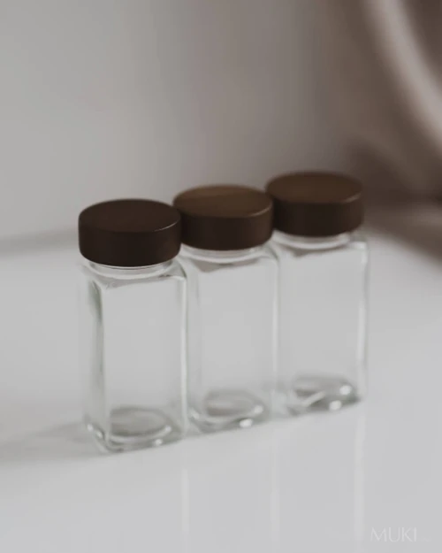 Trois pots à épices carrés en verre vides à couvercles en bois foncé, alignés devant un fond clair