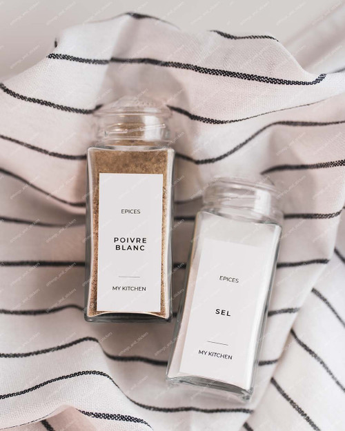 Deux pots à épices carrés en verre couchés sur un torchon rayé blanc et noir, étiquettes Epices Poivre blanc et Sel, vus de dessus
