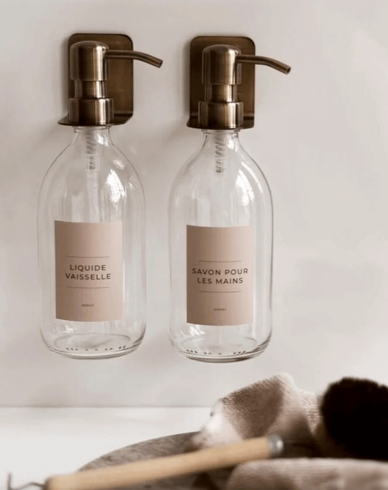 Deux distributeurs de savon muraux en verre transparent sur support bronze, étiquettes Liquide vaisselle et Savon pour les mains