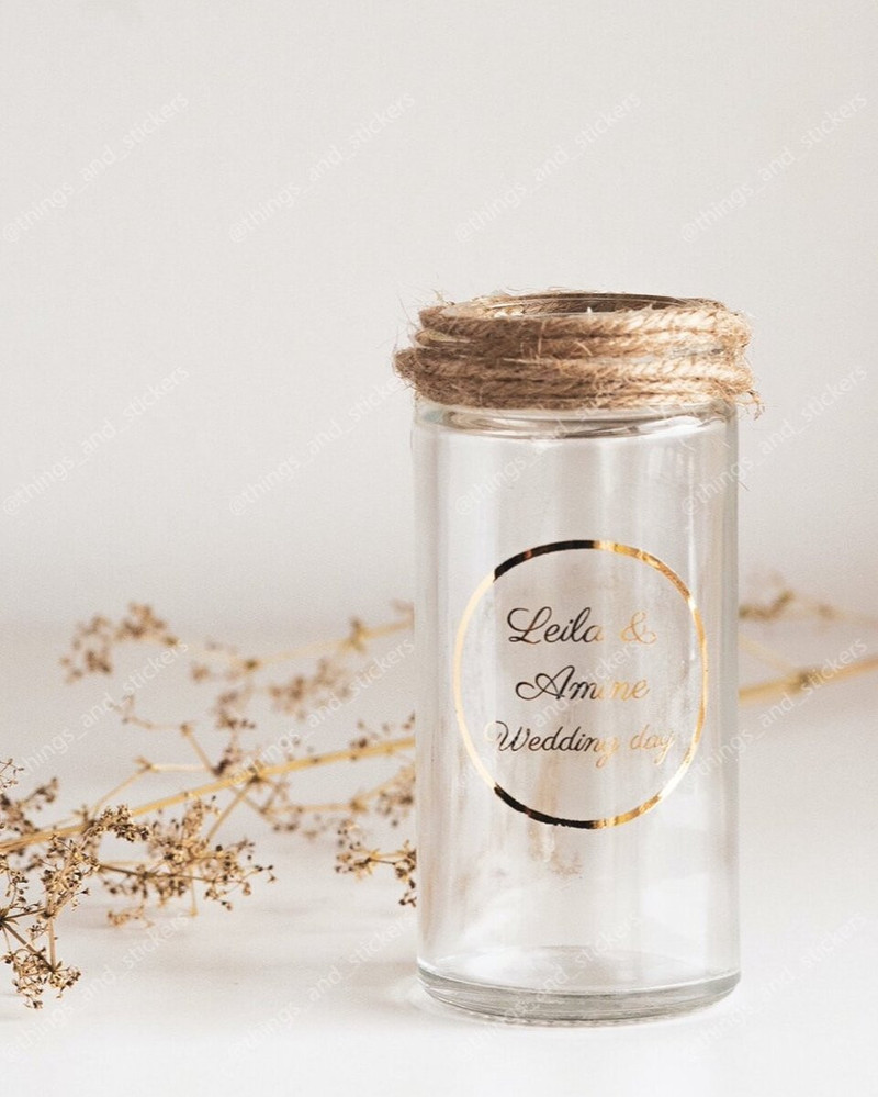 Flacon en verre transparent au col entouré de ficelle, sticker rond doré Leila & Amine Wedding day, près de fleurs séchées