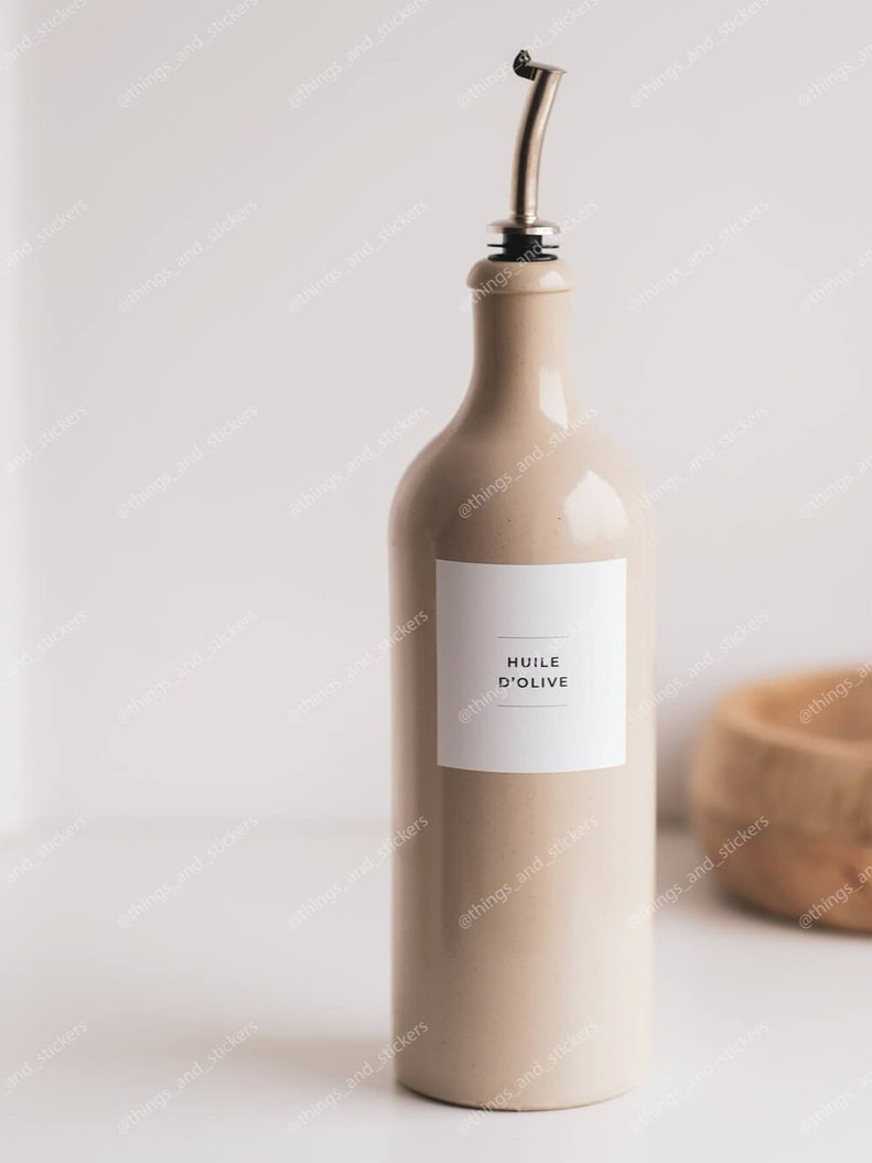 Bouteille huilier en grès beige chaud équipée d'un bec verseur argenté, étiquette Huile d'olive, devant un bol en bois flou