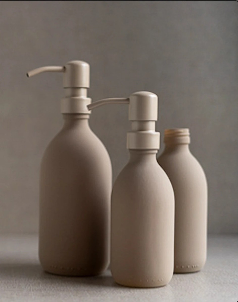 Trois flacons en verre beige dans leurs trois tailles : deux distributeurs à pompe en acier inoxydable beige et un flacon sans pompe, sur fond gris
