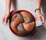 Red dutch oven with marble rye bread. hands lifting the bread of out the Dutch oven using the white long narrow tabs on the round loaf lifter.