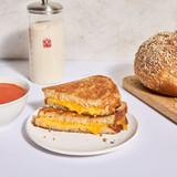 Baker's Grain Sourdough bread recipe made into a grilled cheese sandwich. A loaf of the bread is to the right and a bowl of tomato soup is on the left. The Petite Sourdough Crock sourdough crock is in the background.