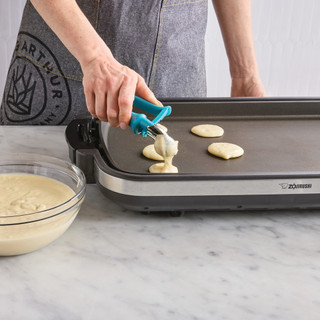 Teaspoon scoop being used to scoop pancake batter onto a griddle.