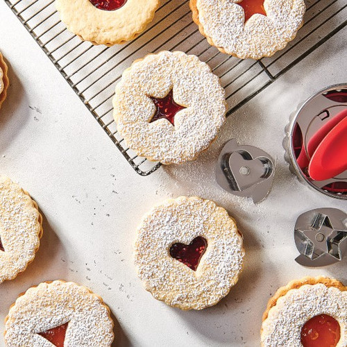 Cardamom Cream Cheese Linzers cookies on cooling rack. Round, light colors cookies, with heart, star, or circle cut out centers with fruit filling.