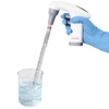 Hand in blue glove holding a white and gray electronic pipette controller with a glass graduated pipette in a beaker.