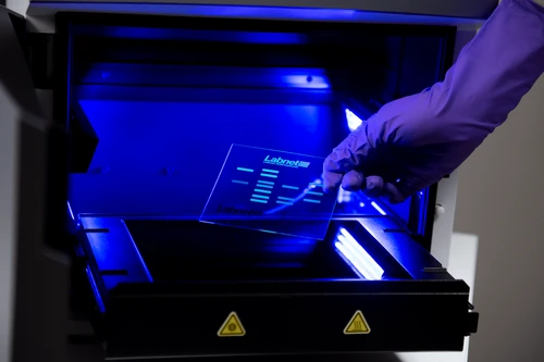 A gloved hand holds a DNA gel sample in front of a blue-lit Labnet gel documentation system.