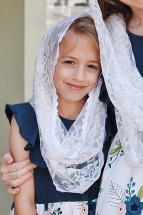 Front view of a young girl wearing the Off-White Girls' Starter Infinity veil, showing the sheer floral lace draped softly over her head and shoulders—a beautiful and comfortable choice for a First Communion veil.
