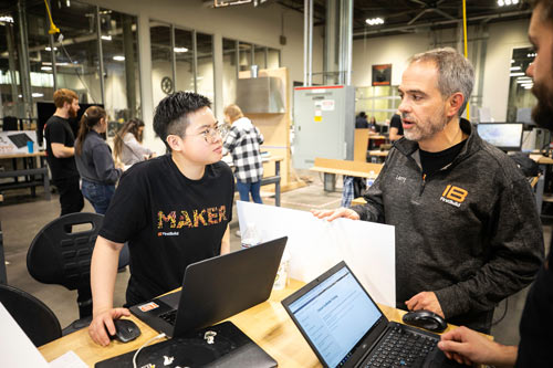 A young Asian man and middle aged white man have a discussion while working at their laptops.