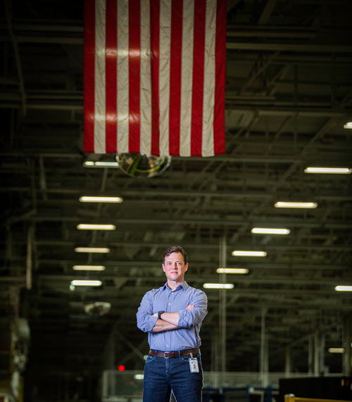 A warehouse worker stands with his arms crossed under an American flag.