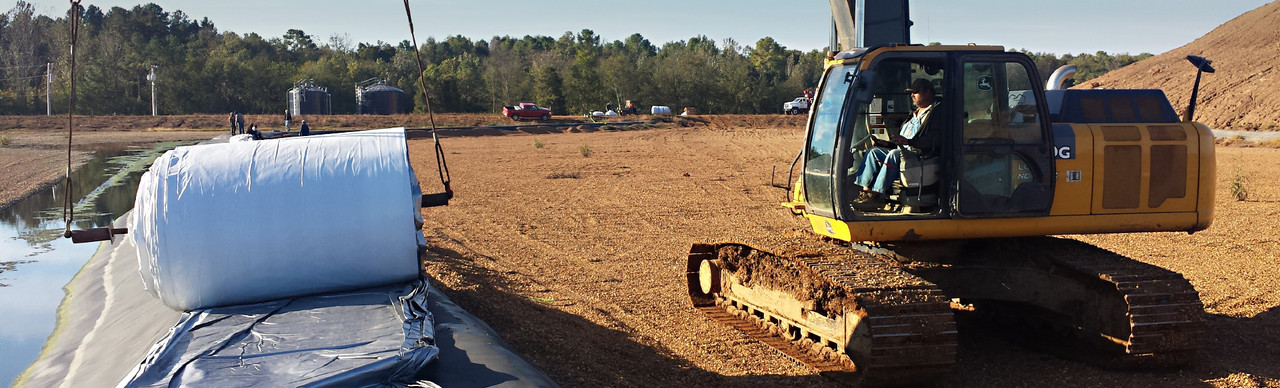 Excavator moves large landfill cover by pond on construction site, with trees and equipment in background.