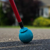 A Desert Blue roller ball tip is attached to an Awarewolf Gear Urban Xplorer cane, shown making contact with a paved surface. The red cane and blue tip are in sharp focus, while the background is softly blurred, suggesting a suburban or urban outdoor environment.