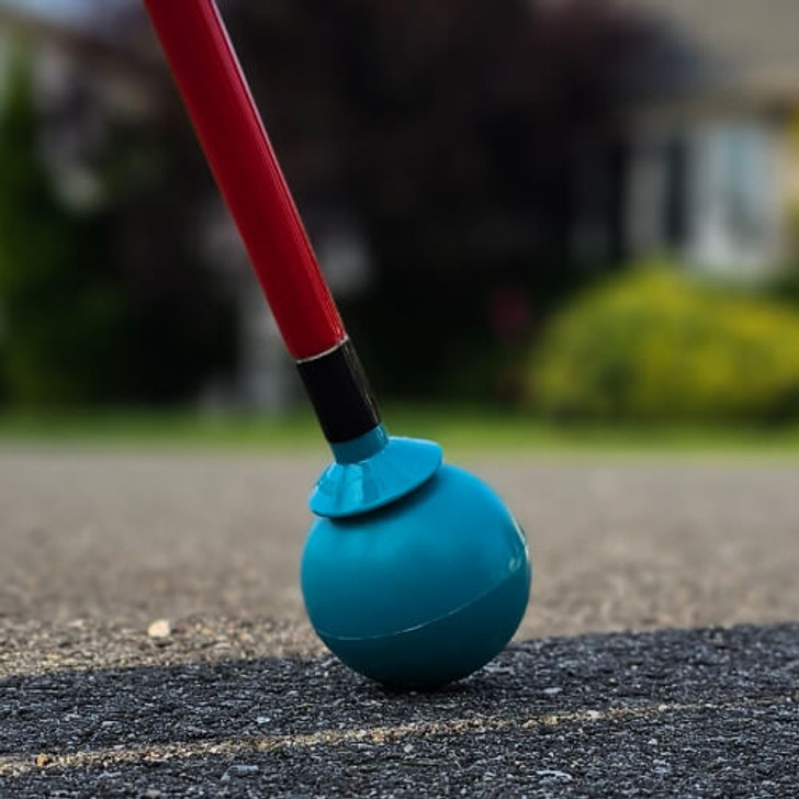 A Desert Blue roller ball tip is attached to an Awarewolf Gear Urban Xplorer cane, shown making contact with a paved surface. The red cane and blue tip are in sharp focus, while the background is softly blurred, suggesting a suburban or urban outdoor environment.