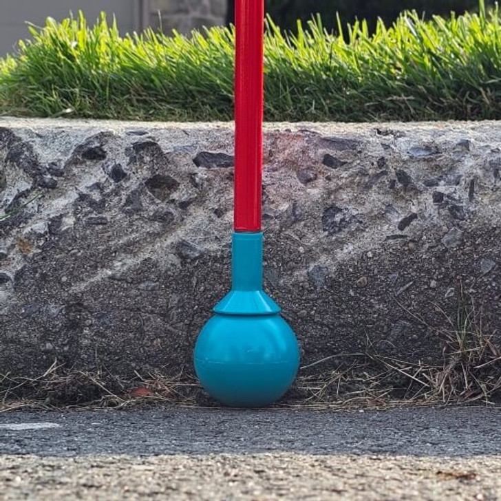 A Desert Blue roller ball tip is attached to an Awarewolf Gear All Terrain Cane, shown making contact with a paved surface. The red cane and blue tip are in sharp focus, while the background is softly blurred, suggesting a suburban or urban outdoor environment. Slip on tip.