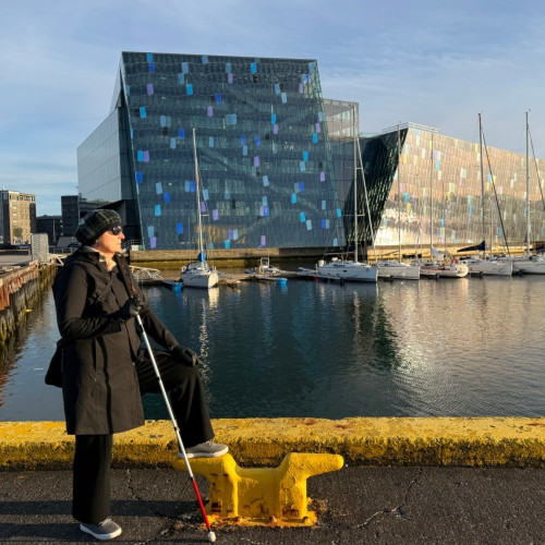 A woman wearing a black coat, hat, and gloves stands confidently by a harbor holding an Awarewolf Gear **Urban Xplorer Cane**. Sailboats and a modern glass building shimmer in the background, symbolizing independence, style, and exploration with the Awarewolf Gear × NoisyVision collaboration.
