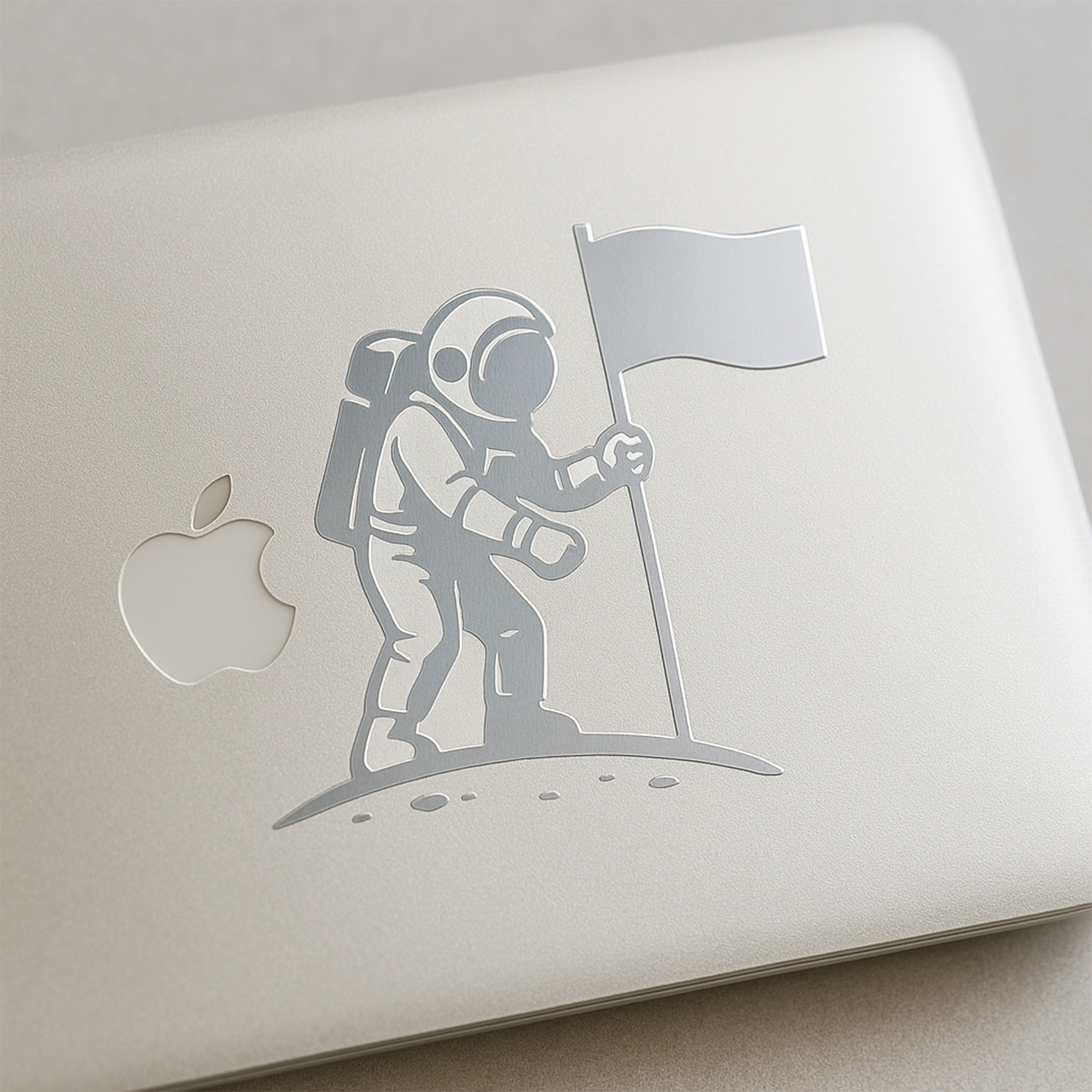 Minimalist silver vinyl decal of an astronaut planting a flag, applied to a silver MacBook.