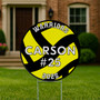 Round volleyball-themed yard sign with yellow and black design, the word "WARRIORS" at the top, name "CARSON" in bold white letters, jersey number #25 in the center, and "2025" at the bottom, displayed on a wire stand in a residential lawn.