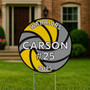 Close-up of the personalized volleyball yard sign staked into grass with a blurred park-like background. The design includes “WARRIORS” arched across the top, bold “CARSON” name across the middle, “#25” centered, and graduation year “2025” curved along the bottom—all layered over a yellow and gray volleyball graphic.